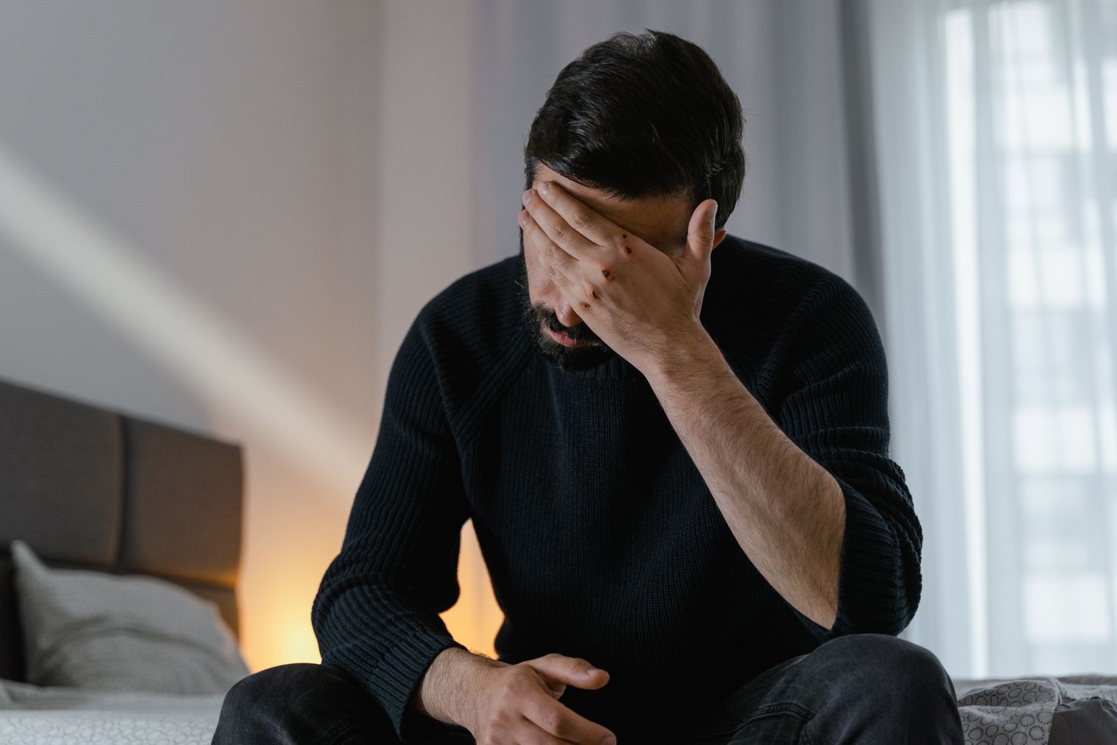 A man in distress sits on a bed indoors, covering his face with his hand.