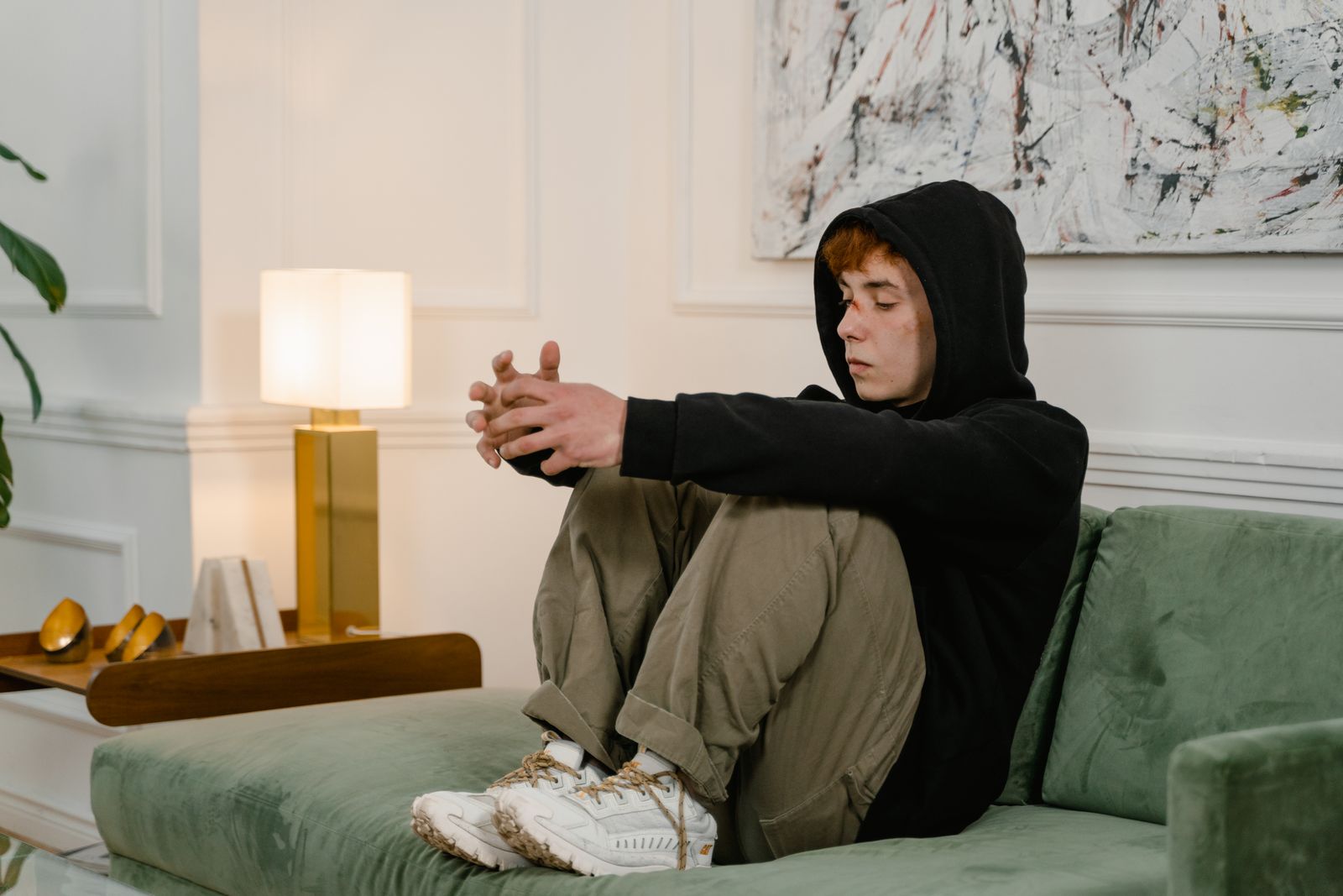 A young man in a black hoodie sits thoughtfully on a green couch indoors.