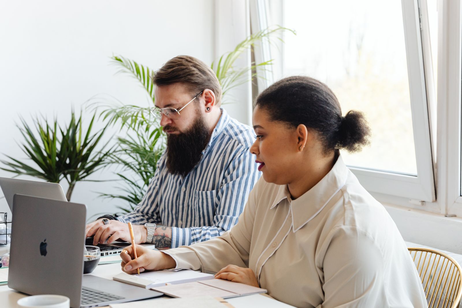 Two colleagues working together in a bright modern office space with laptops and notepads.