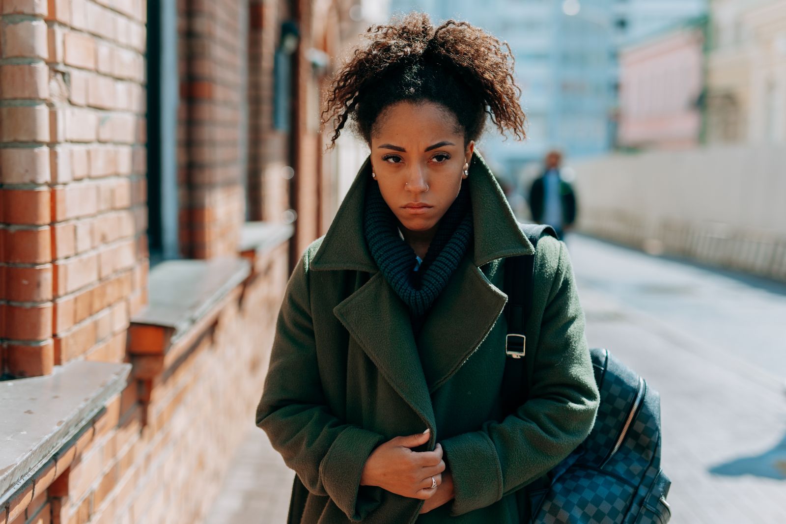 A thoughtful woman in a green coat walks along a sunlit urban sidewalk, lost in contemplation.