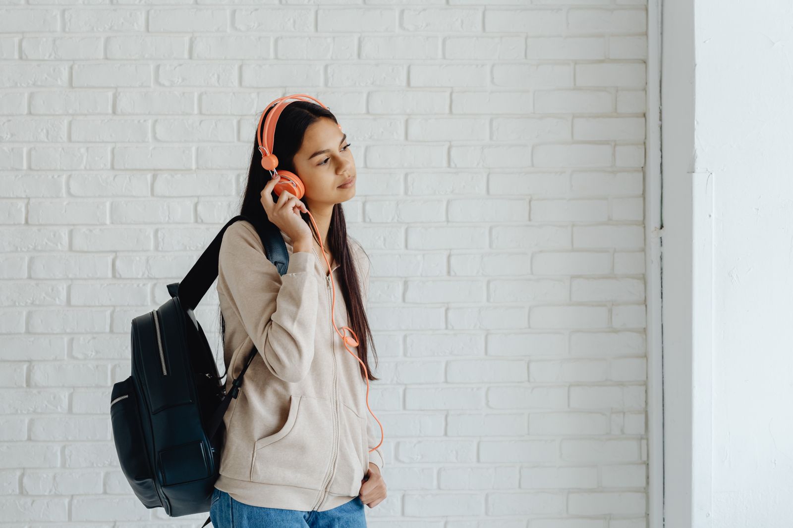 Teen girl with headphones standing against a white brick wall indoors, lost in music.