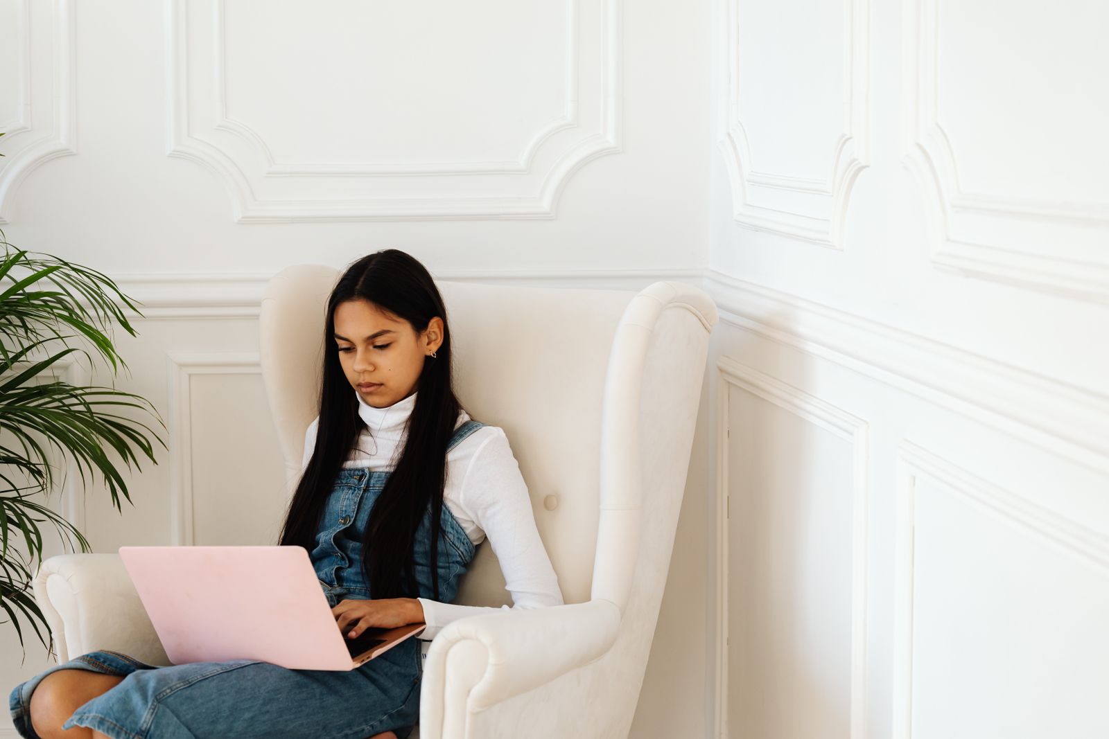 Teen girl in denim dress using a pink laptop while sitting on a white armchair indoors.