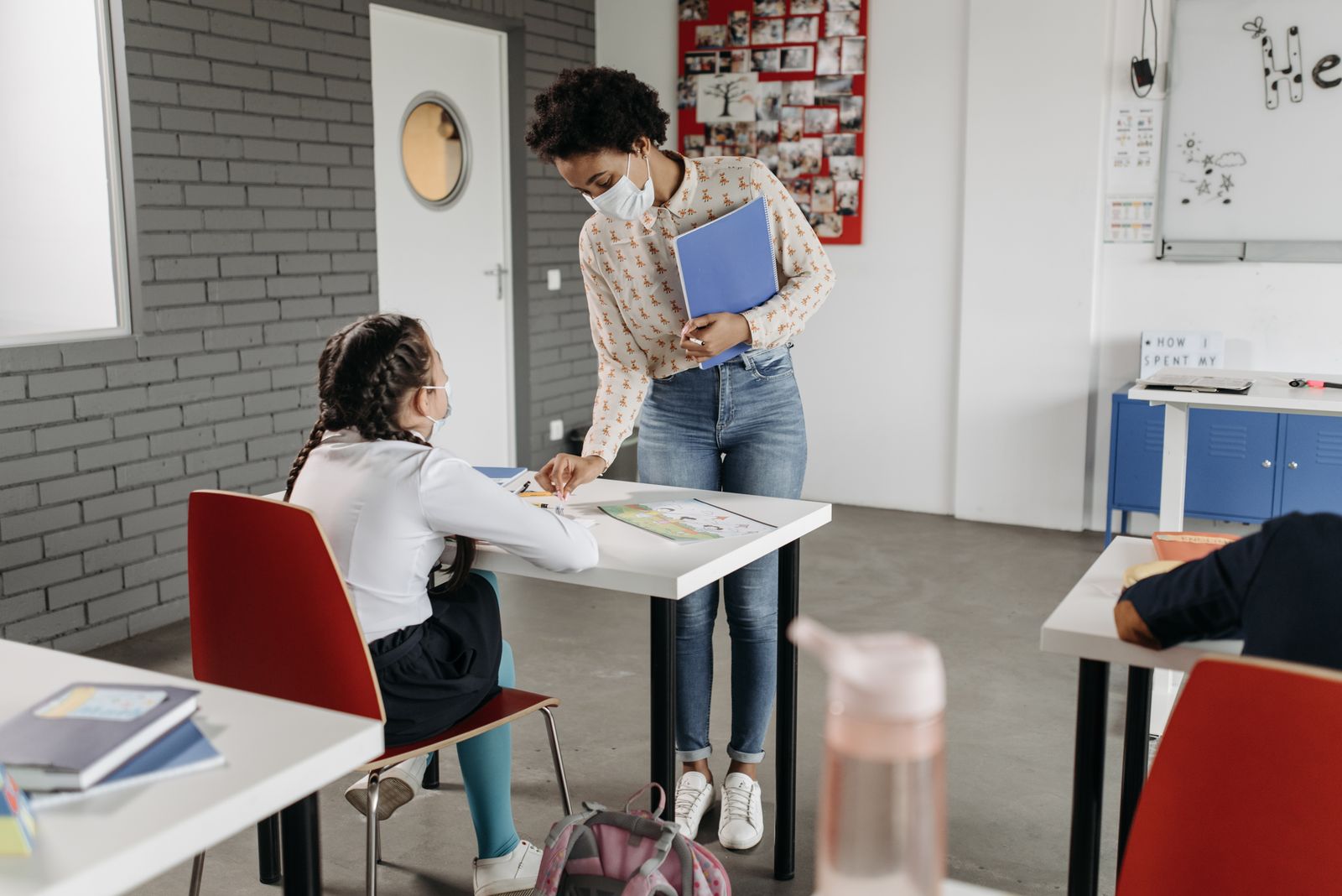 Teacher assisting a student in a classroom while both wear face masks, following safety protocols.