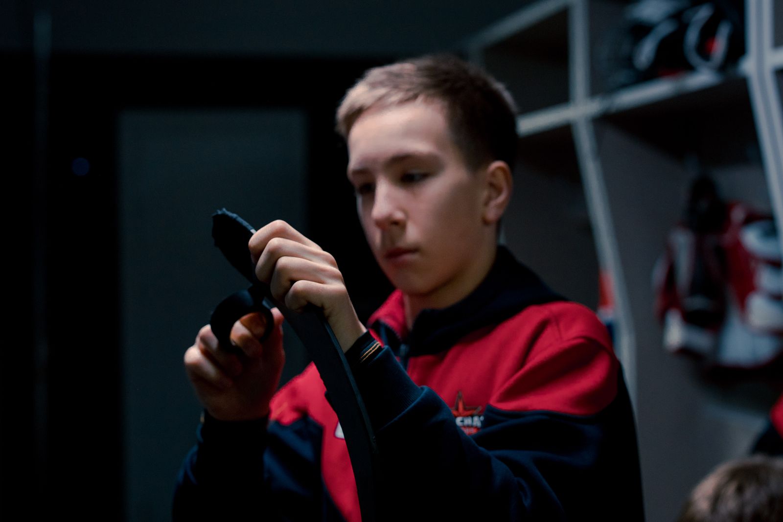 Teenager in sports jacket taping hockey stick in indoor locker room setting.