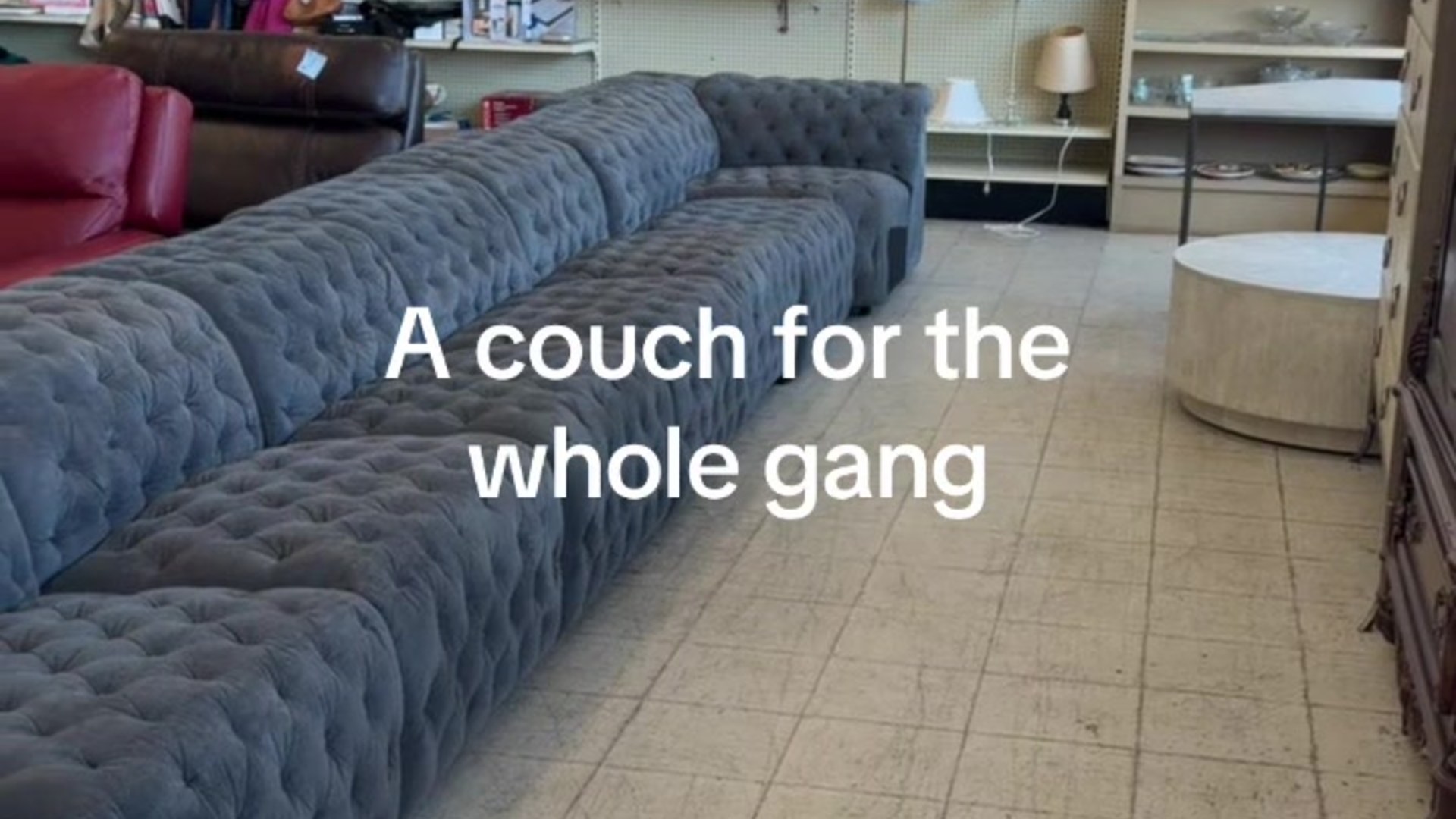 A very long grey tufted sectional couch on a thrift store floor with the caption a couch for the whole gang