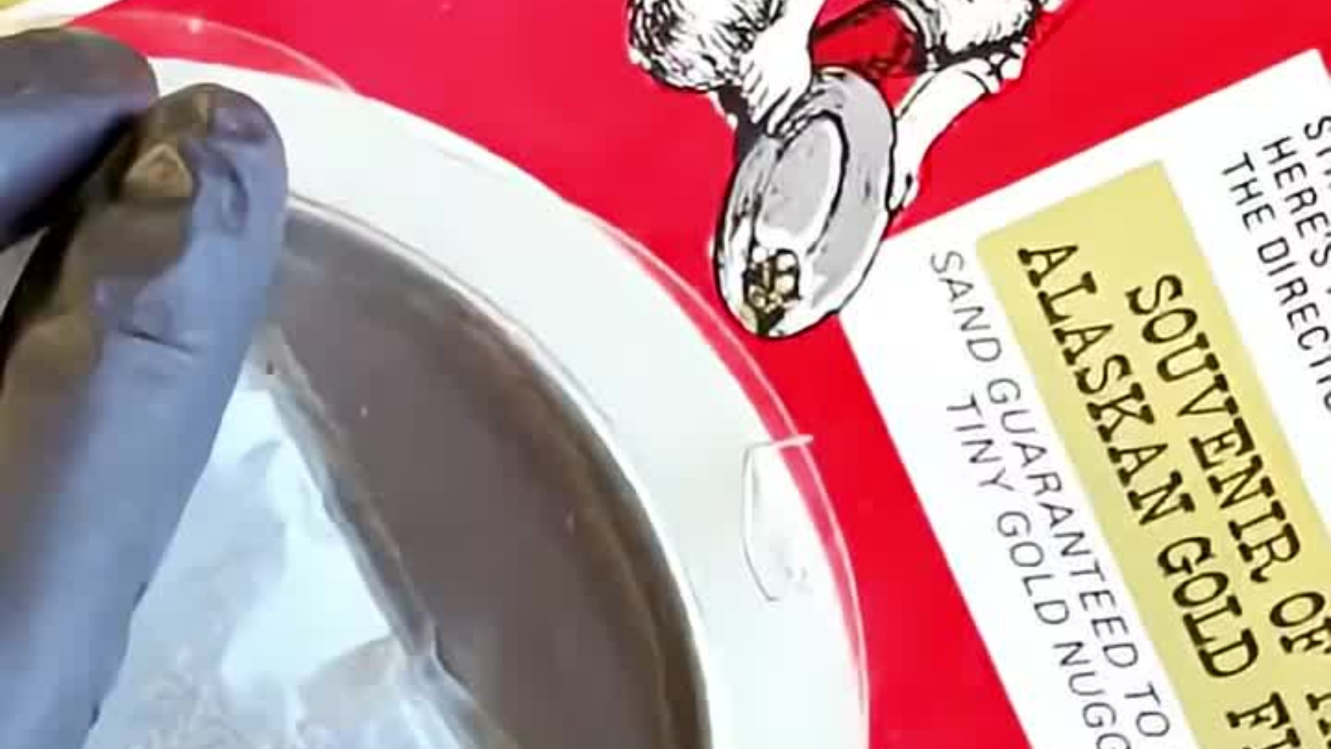 Close-up of a gloved hand over a gold pan beside the red 1972 Souvenir of Alaskan Gold Fields paydirt bag, from the @kleshofficial TikTok