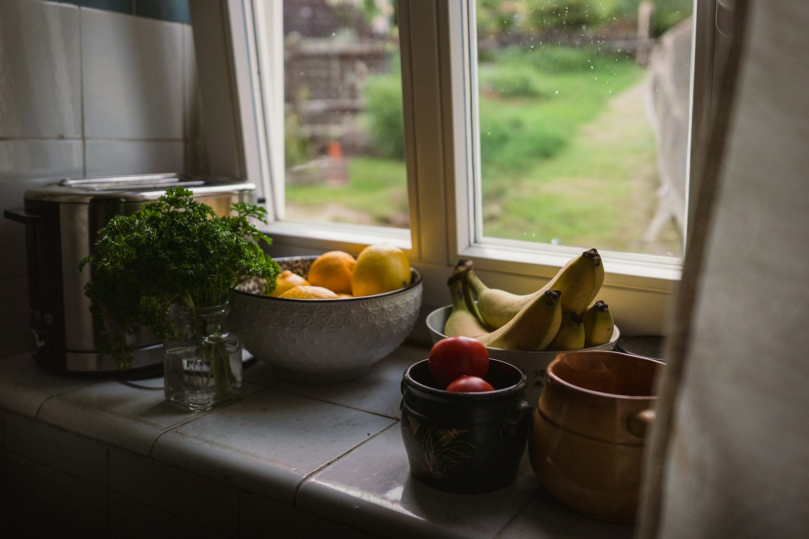 A bowl of lemons, bananas, and tomatoes on a kitchen windowsill with parsley