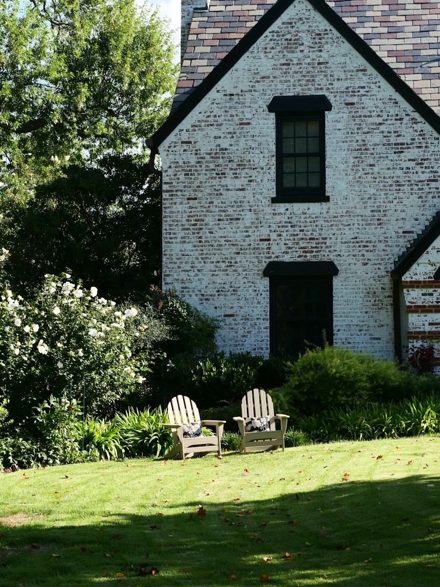 Two wooden chairs on a green lawn beside a white brick house